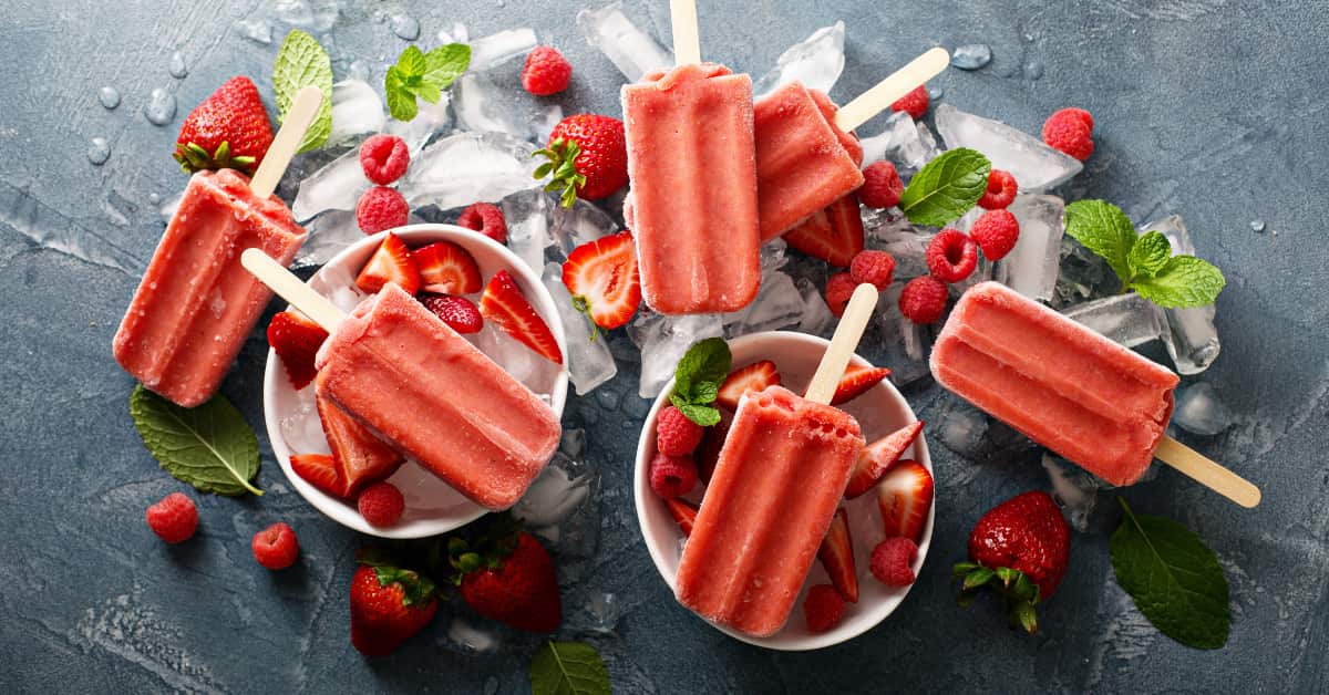 red popsicles with wooden sticks sitting on bowls of sliced strawberries