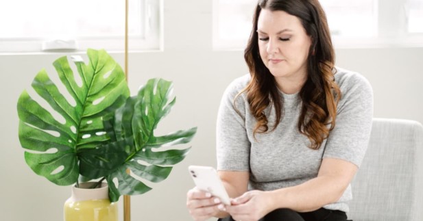 woman sitting on chair next to plant checking cell phone.