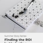 black and white journal and pen with text "summer story series finding the roi in marketing".