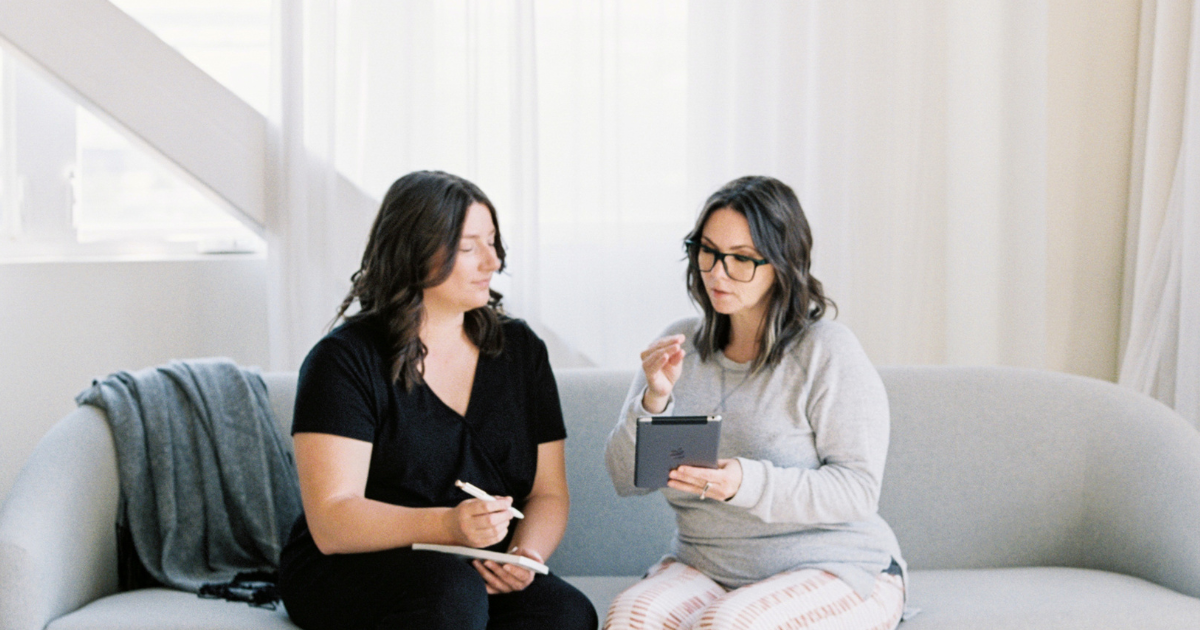 Two women sitting on a couch looking at an ipad and talking.