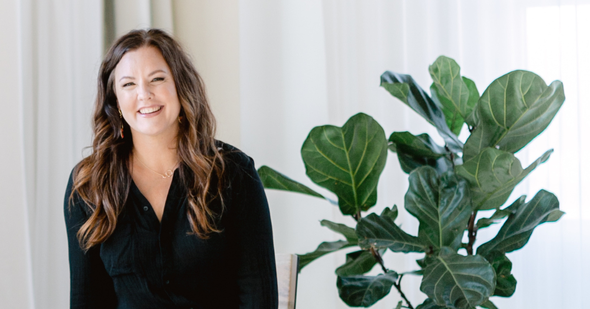 a brunette woman sitting next to a plant, smiling.